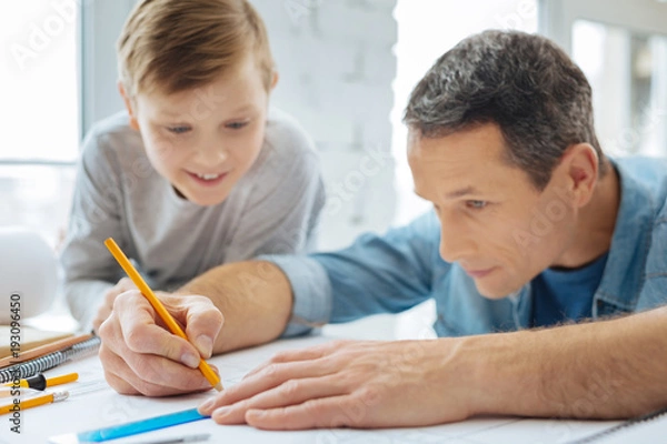 Fototapeta Fascinated by work. Pleasant curious pre-teen boy sitting at the table next to his father and watching him draw a blueprint with the pencil and ruler