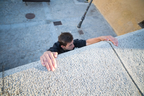 Fototapeta Young man hanging on wall on hands and trying to climb up while doing parkour.  