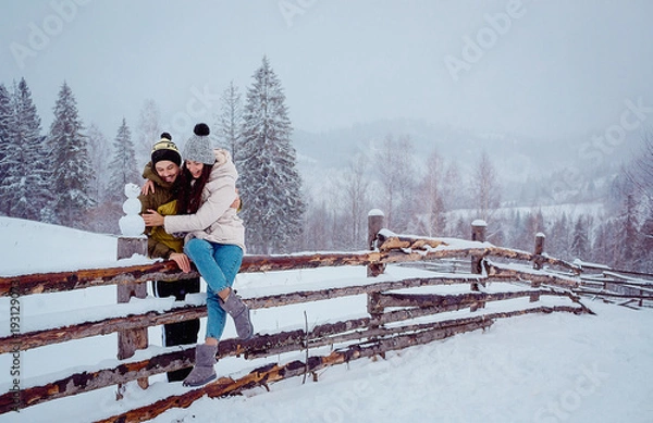 Fototapeta Lovely couple looks funny sitting on the fence covered with snow and with snowman by their side
