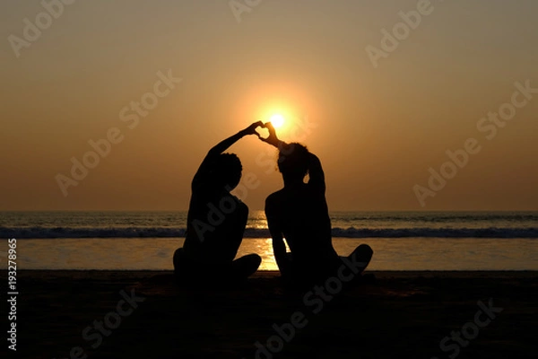 Fototapeta Couple's silhouette drawing heart with their hands on the beach at sunset