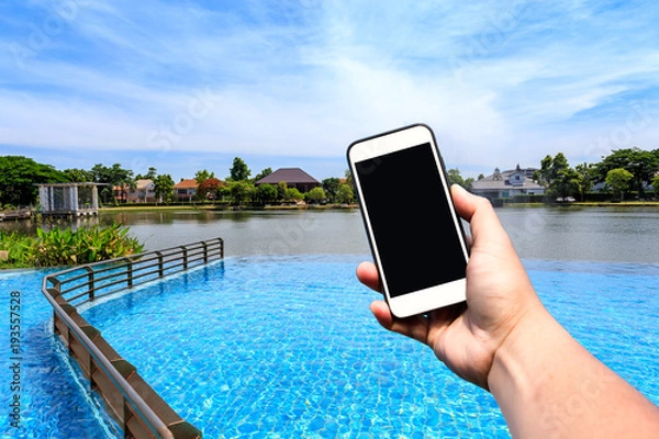 Fototapeta Hand holding blank screen smartphone with beautiful sky and outdoor swimming pool and lake with copy space.