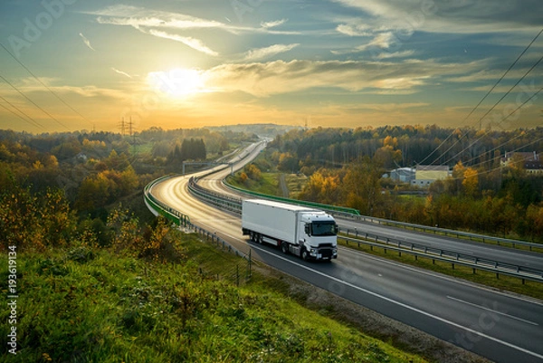 Fototapeta Biała ciężarówka jadąca autostradą wijącą się przez zalesiony krajobraz w jesiennych kolorach o zachodzie słońca