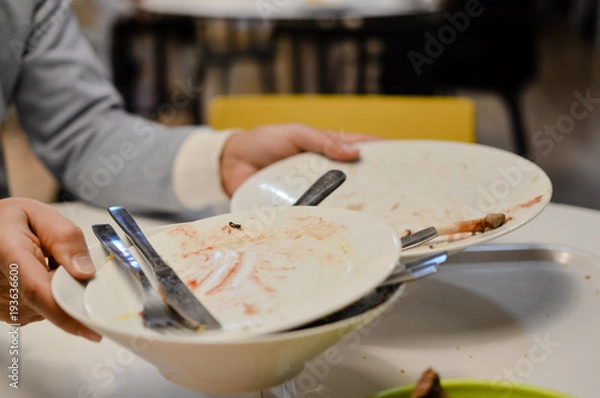 Fototapeta Close up on hand of a man taking dirty dishes and tableware on the finished dinner table background. Eating done clean time, abstract window bokeh space