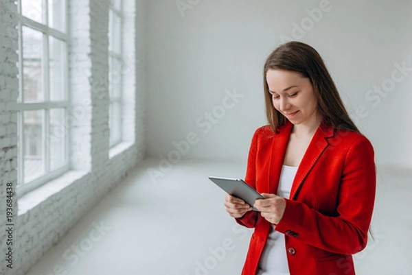 Obraz Portrait of smiling successful adult girl in red jacket looking at her tablet