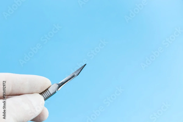 Fototapeta Scalpel in doctors hands in white medical gloves isolated on blue background copy space