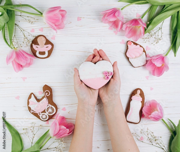 Obraz Female hands holding heart. A composition for newborns on a wooden white background with pink tulips, hearts and a cookies, copy space and flat lay. It's a girl.