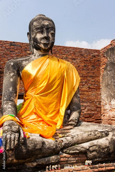 Fototapeta Old Buddha statue in festive safran clothes sitting in lotus position in ruined Wat in Ayuttaya of Thailand