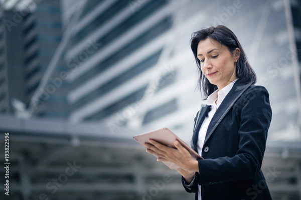 Obraz Caucasian businesswoman with wireless tablet standing outdoors with office building in the background, low angle view
