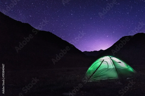 Fototapeta Astrophotography of night camp and Milky way. Dark night and bright galaxy above Caucasus mountains in Georgia. Green tent on the foreground is highlighted from the inside. Backpacking lifestyle.