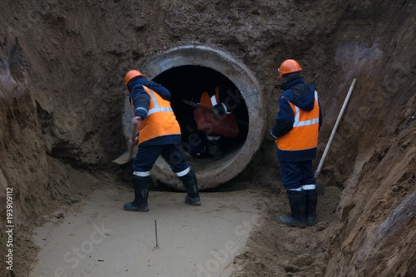 Fototapeta Working under the ground with a large concrete pipe . wet clay. Workers in overalls, helmet, protective boots. A deep excavation .