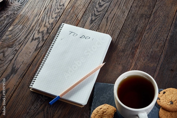 Fototapeta Cup of tea with cookies, workbook and a pencil on a wooden background, top view