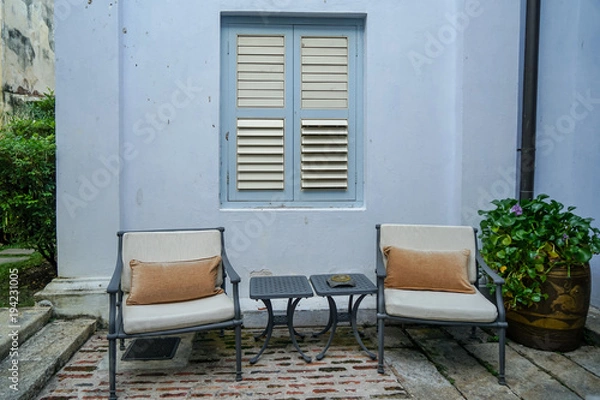 Fototapeta Outdoor garden furniture, grey metal lounge chairs and side tables in front of blue building wall with traditional window on concrete floor next to water plant in local pot, Penang