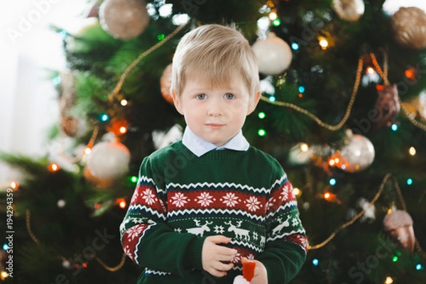 Fototapeta Pretty little boy in green sweater stands with a toy before a Christmas tree