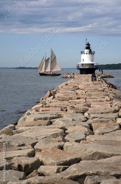 Fototapeta Schooner Sailboat Passes by Lighthouse Situated on Edge of Stone Jetty in Maine