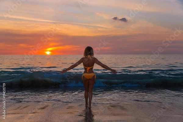 Obraz красивая женщина с шикарной фигурой в оранжевом бикини стоит спиной к фотографу и лицом к морю на фоне заката 