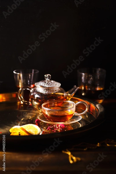 Obraz Small glass teapot and glasses with hot black tea, dried rose petals, pocket magnifier on golden chain, squeezed orange slice on golden tray. Evening light. Dark background
