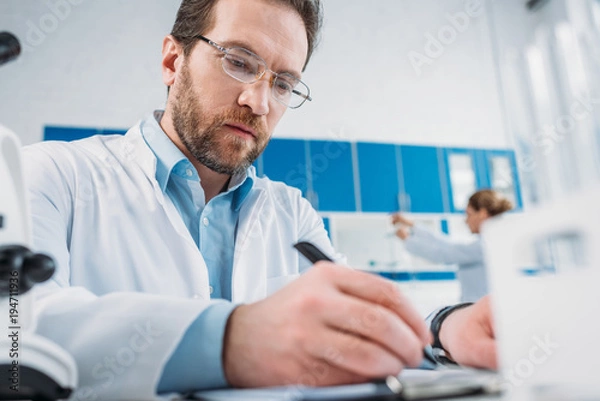 Obraz selective focus of scientist in white coat and eyeglasses making notes in notepad at workplace in laboratory