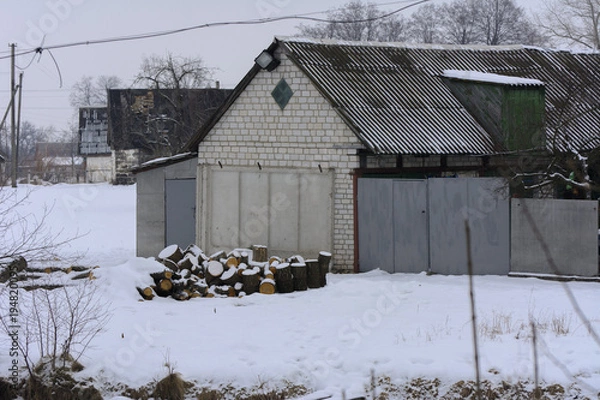Fototapeta A village dwelling house with outbuildings fenced with a stone fence against a background of white snow in the winter.