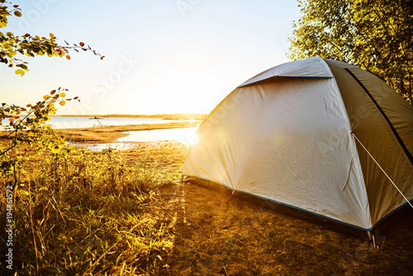 Obraz White tent standing at a beach camping spot at lake Vänern in Sweden. The sun is shining and soon will be sunset. Tent is standing on the right side covered by trees  and plants. Water reflections.  