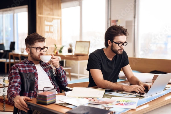Obraz Two freelancer men working at laptop and drinking coffee at desk.