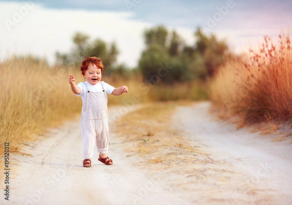 Fototapeta adorable redhead toddler baby boy in jumpsuit running along rural summer road in sunburned field