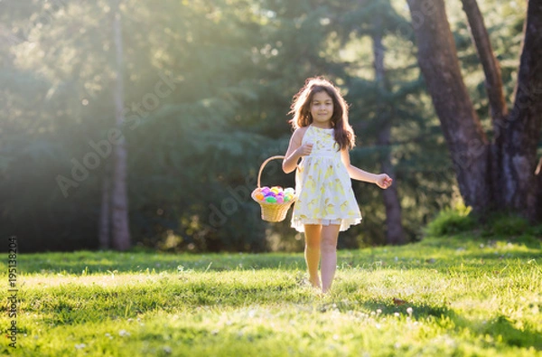Fototapeta Little girl walking on green grass toward camera holding woven basket with pastel Easter eggs