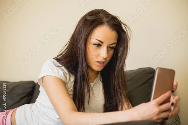 Obraz Beautiful woman in casual clothes lying on leather sofa and using smartphone