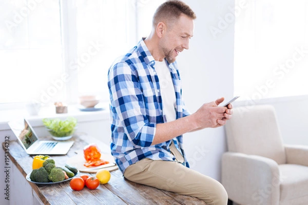 Fototapeta Biohackers recipes. Joyful merry jolly man using phone for looking through article about biohacking and sitting on surface