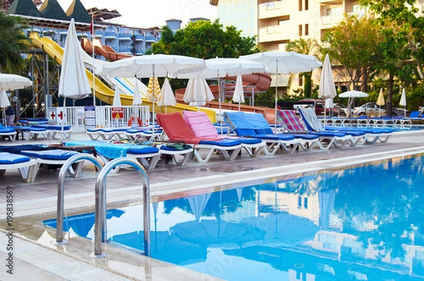 Fototapeta Swimming pool, sun loungers with towels, beach umbrellas, but no people. A typical summer vacation is all inclusive. A hot summer day. Water slides, mini water park in the background.
