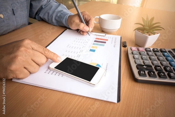 Fototapeta Business man analyzing investment charts with smart phone and calculator on office desk table.Close up.Financial Report Revenue Statistical Accounting Concept.