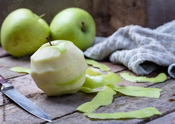 Fototapeta Green peeled apple on wooden desk with knife and apples behind.