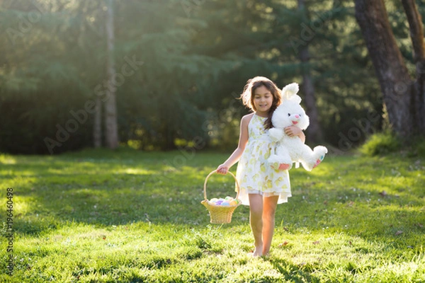 Fototapeta Little girl walking barefoot toward camera, holding Easter basket with eggs and plush toy big bunny