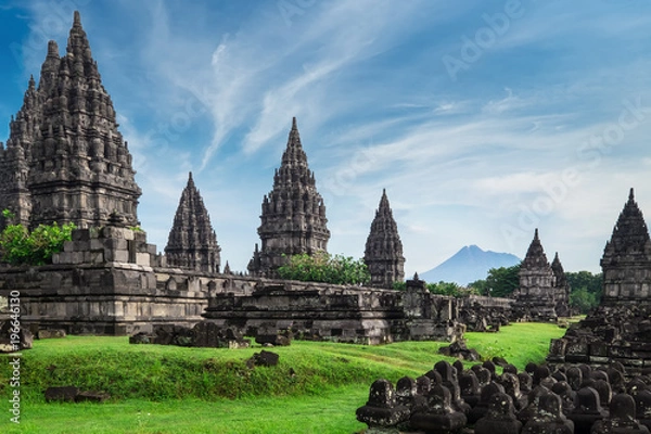 Fototapeta Ancient stone ruins on green field and Candi Prambanan or Rara Jonggrang, Hindu temple compound on background. Impressive architectural site. Yogyakarta, Central Java, Indonesia. Panoramic view.