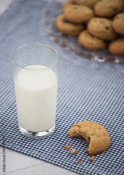 Fototapeta A glass of milk and a cookie with a bite taken out of it sit on a blue gingham napkin with a plate of cookies in the background