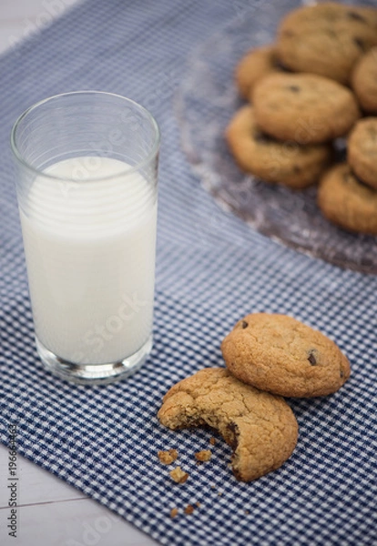 Fototapeta A glass of milk and a cookie with a bite taken out of it on a blue gingham napkin with a plate of cookies in the background