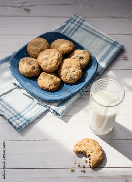 Fototapeta A glass of milk and a cookie with a bite taken out of it next to a blue plaid napkin with a plate of cookies in the background