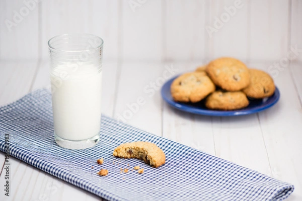 Fototapeta A glass of milk and a cookie with a bite taken out of it on a blue plaid napkin with a plate of cookies in the background