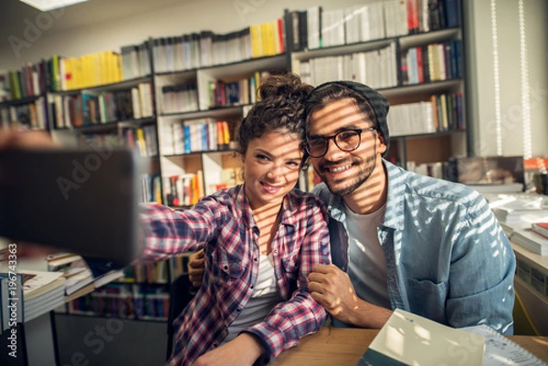 Fototapeta Close up of smiling hipster cheerful young student love couple sitting in the high school library and taking a selfie near the sunny window.