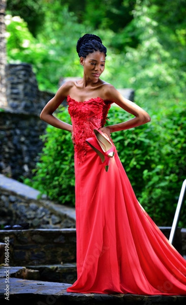 Obraz African American girl in a red dress, with dreadlocks, with red shoes in hand, posing in the summer in the Park on a background of green plants. Portrait. Vertical view