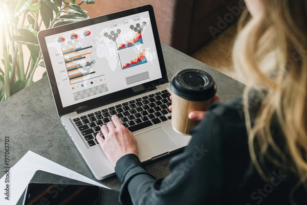 Obraz Rear view.Young businesswoman is standing near table,working on laptop with graphs, charts,diagrams, schedules on screen