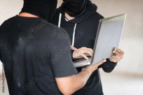 Fototapeta Two black masked hackers are using laptop in them hand. They are trying to  hack data. Selective focus at far hacker' hand.