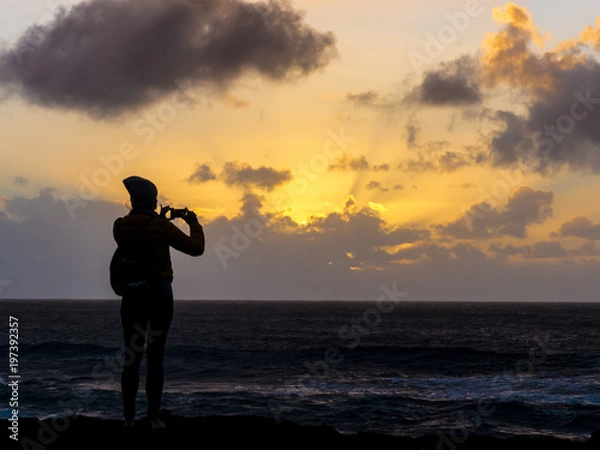 Obraz Girl standing at the Atlantic coast captering the sunset with a smartphone.