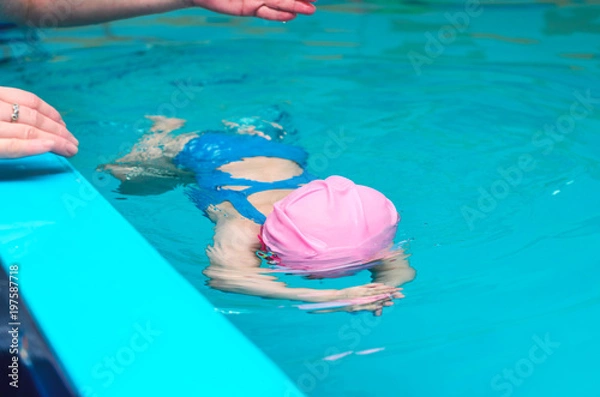 Fototapeta A little girl of European appearance in a pink rubber cap learning to swim in the pool in glasses for swimming