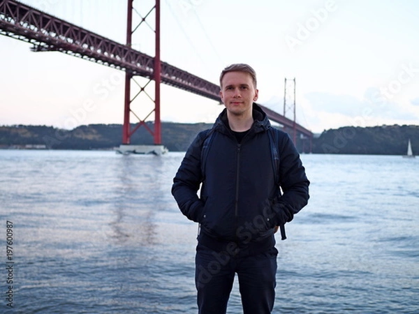 Fototapeta A man in a jacket stands against the background of the sea city and the bridge in  Lisbon.