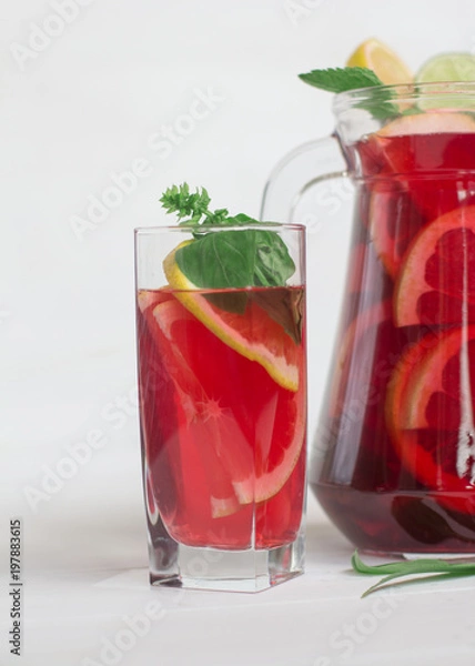 Fototapeta A glass with red sangria and oranges, lime and lemon on a white background. Behind the jug.Close up. Copy-space.