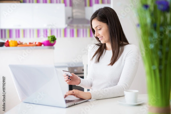 Fototapeta Young beautiful happy girl is typing her credit card info on a laptop while drinking coffee and sitting at the table of bright kitchen.
