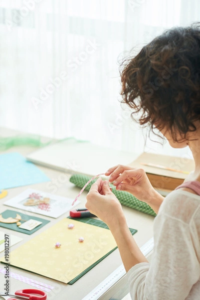 Fototapeta Profile view of curly woman in casualwear sitting at wooden table while enjoying process of handmade greeting card creation