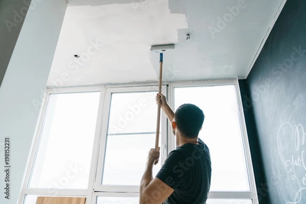 Fototapeta the man paints the walls and the ceiling in gray color, standing with his back to the camera. Focus on the roller. Painting and repair of the room.