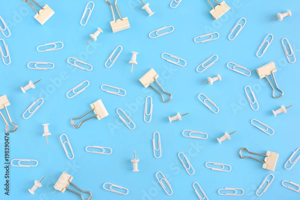 Fototapeta Stationery chaotically scattered on a blue background. White paper clips and clerical buttons in the riots. Photo from the top.