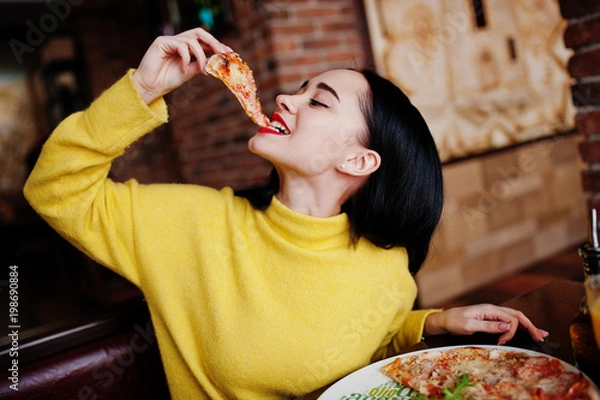 Fototapeta Zabawna brunetka dziewczyna w żółtym swetrze jedzenie pizzy w restauracji.